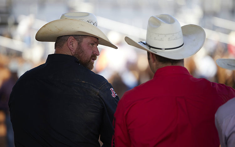 A cowboy wearing a black shirt looking at a cowboy wearing a red shirt at a rodeo.